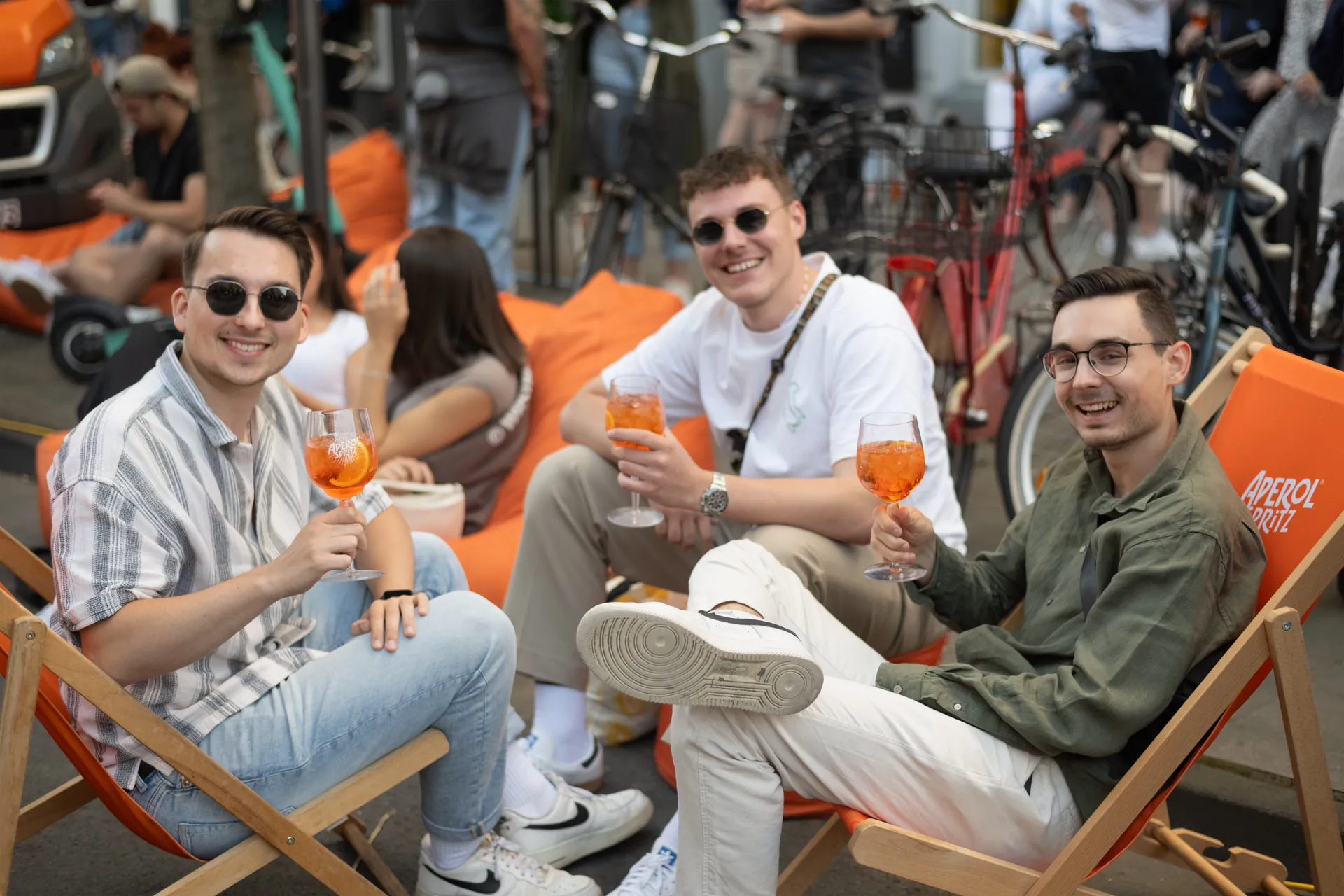 Drei lachelnde junge Manner sitzen in orangefarbenen Stuhlen und halten Aperol-Spritz-Glaser in der Hand.