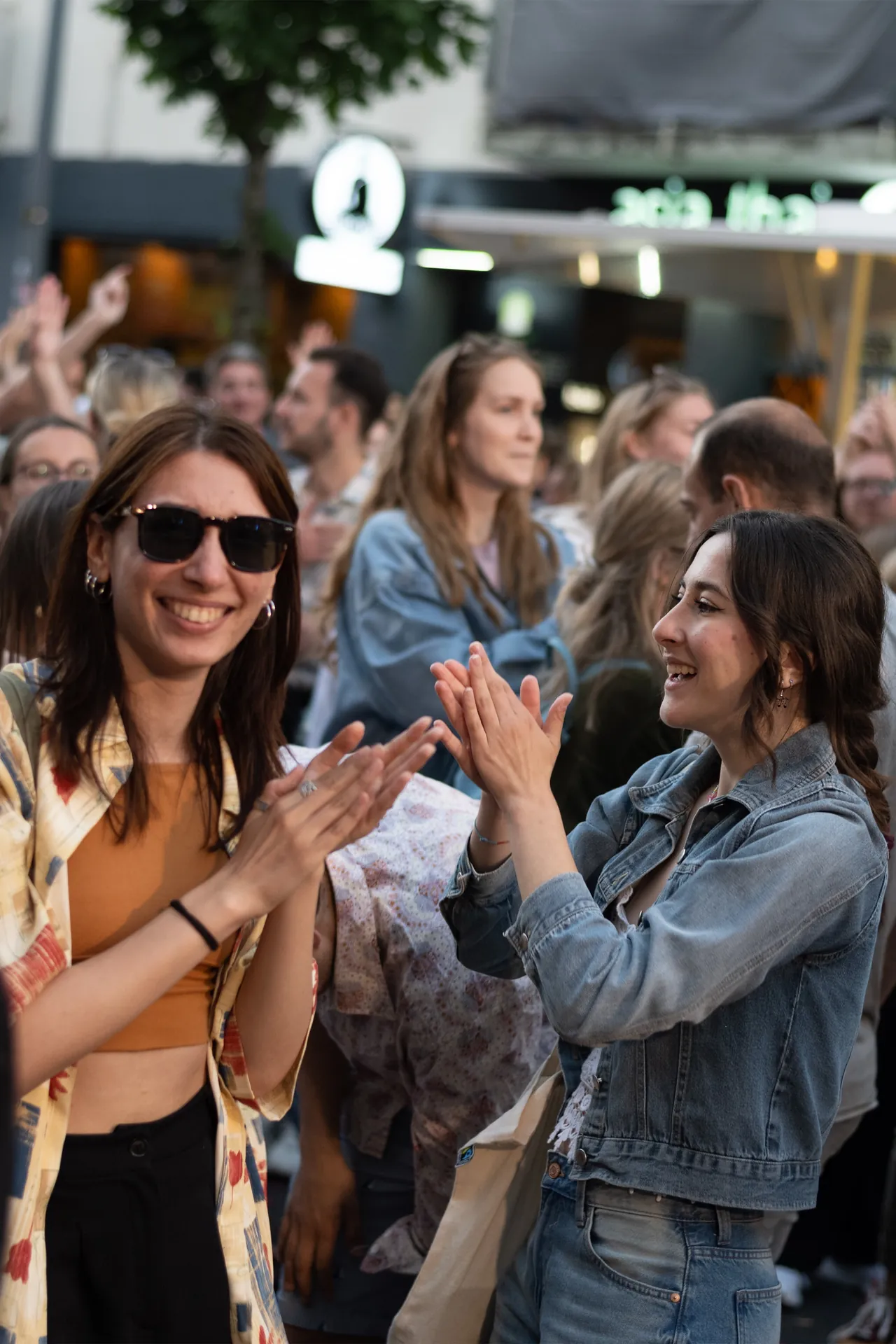 Zwei junge, lächelnde Frauen klatschen im Publikum, weitere Gäste im Hintergrund, Bonner Straße im Bild sichtbar.