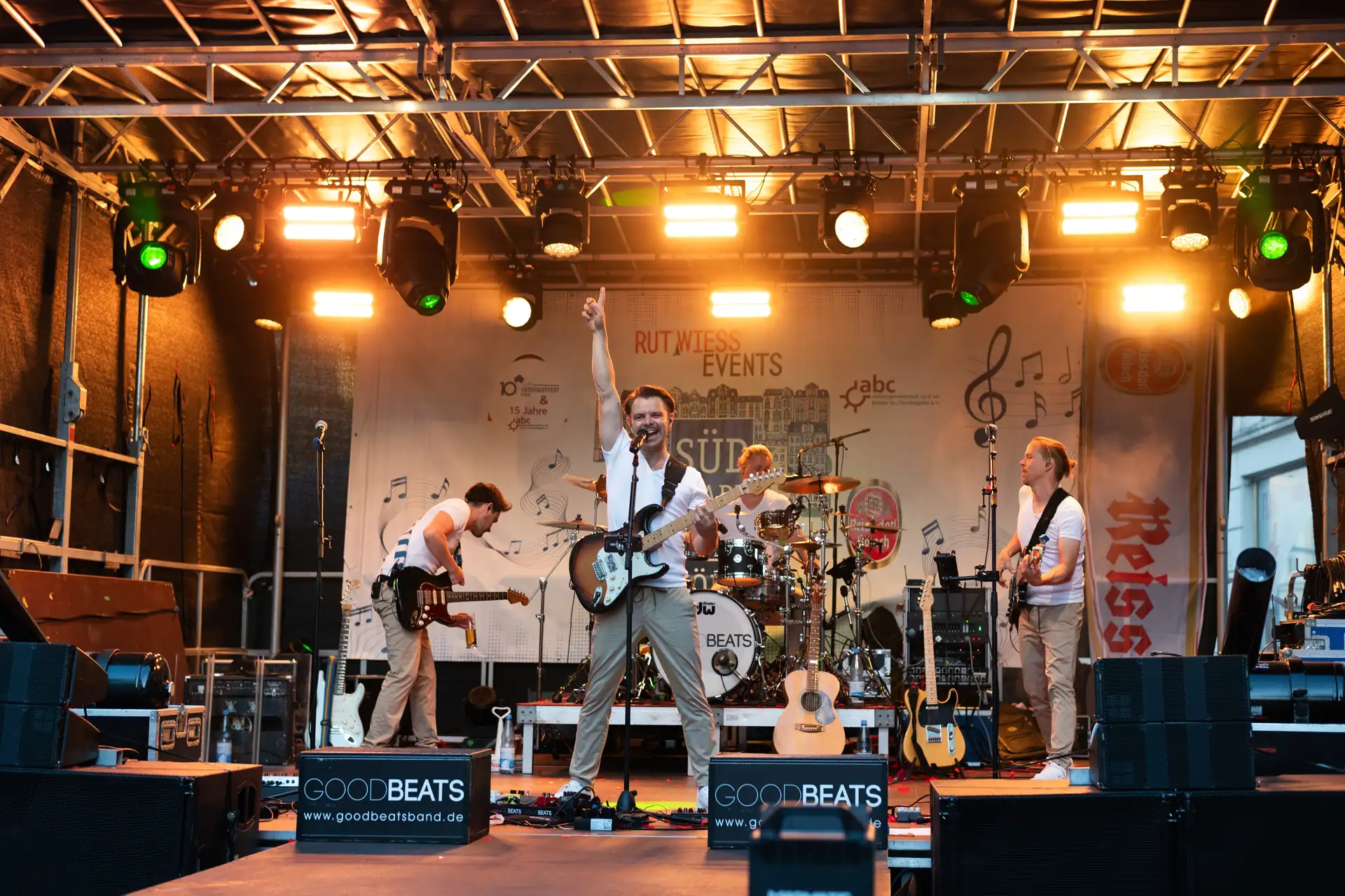 Eine Band spielt mit Gitarren und Schlagzeug auf einer Bühne mit Lichteffekten und Bannern.