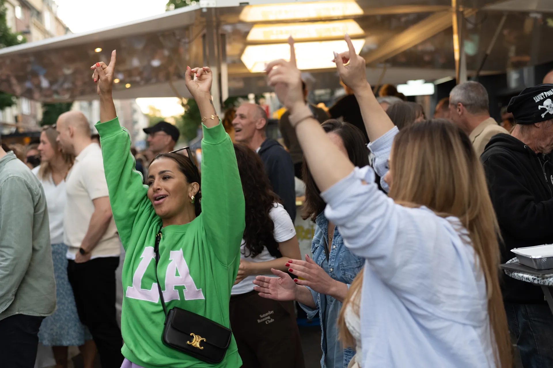 Eine ausgelassene Menschenmenge feiert auf einem Event, die Arme hochgerissen, eine junge Frau im Vordergrund mit einem grünen Pullover.