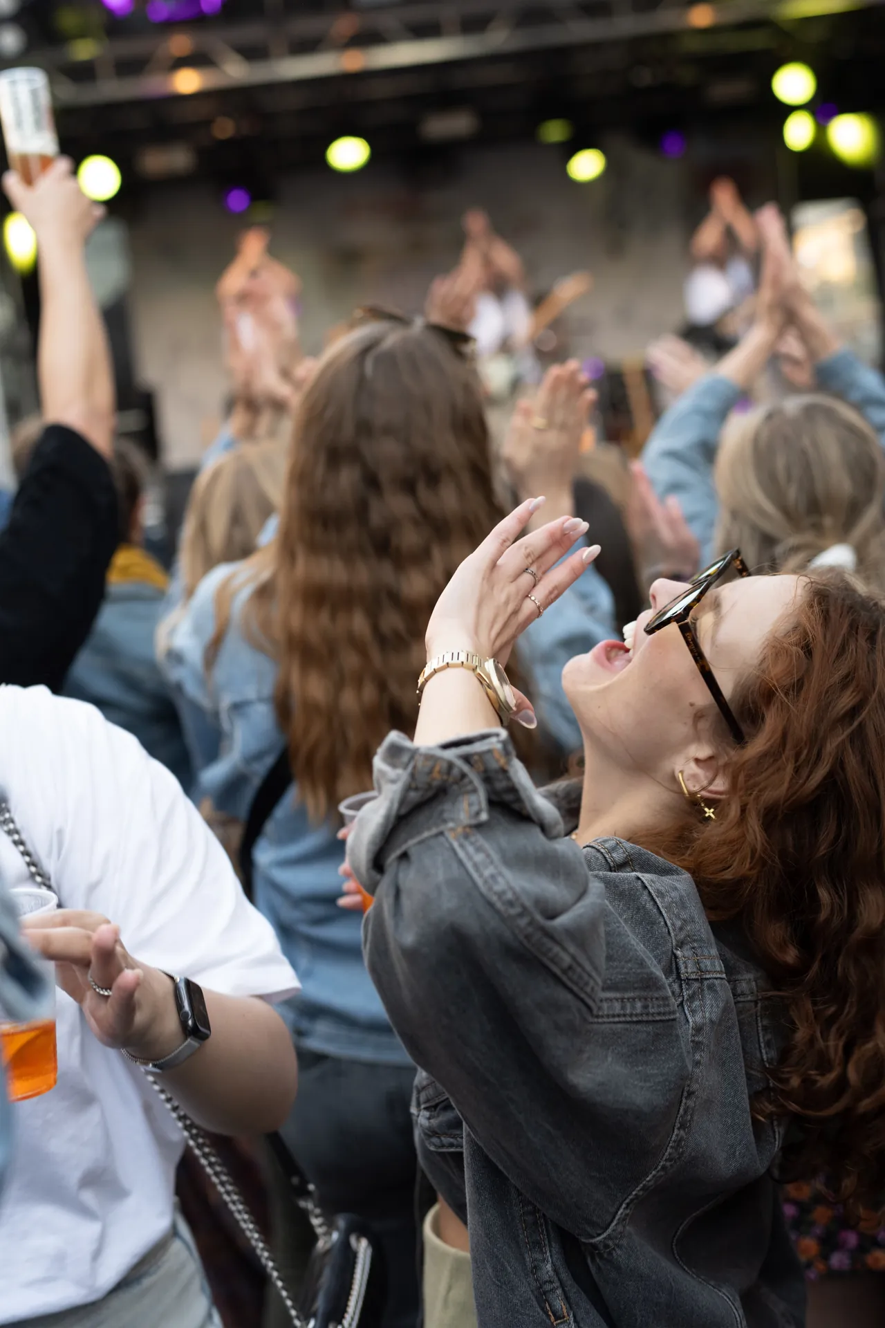 Eine lachende Frau mit lockigen, roten Haaren feiert auf einem Event mit weiteren Gästen vor einer Bühne.