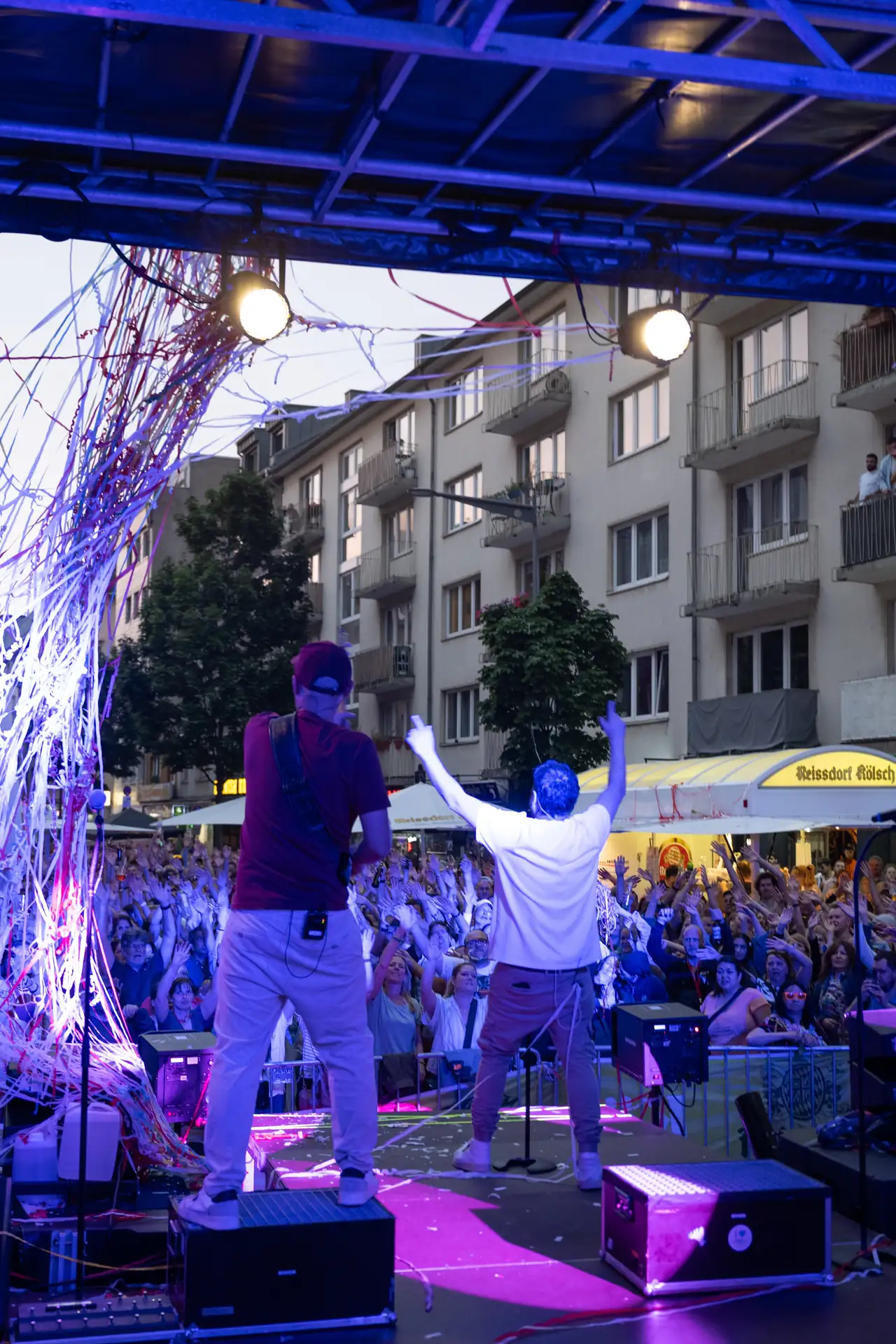 Zwei Musiker stehen auf einer Bühne vor begeistertem Publikum, das die Hände in die Luft wirft.