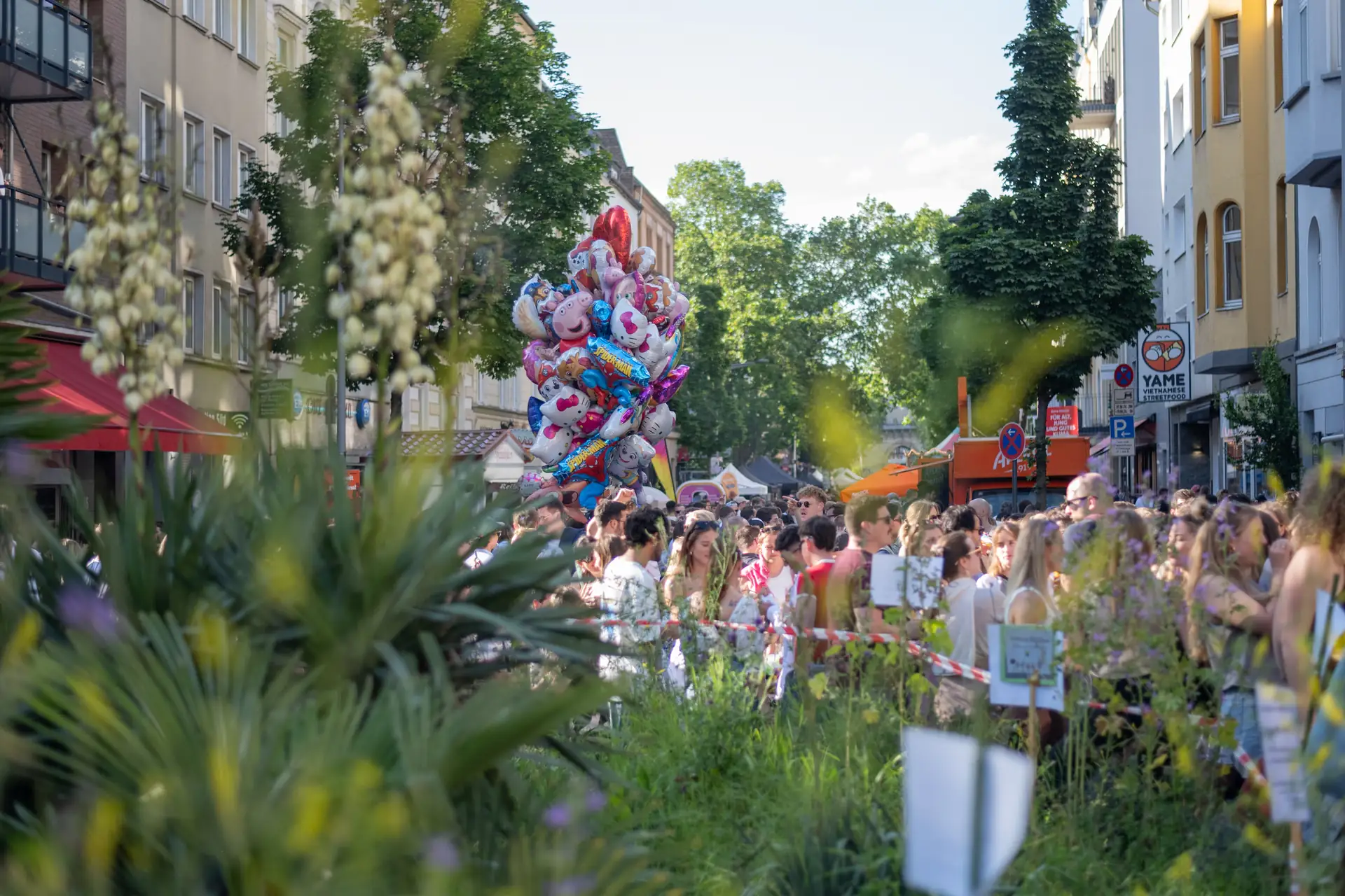 Eine grosse Menschenmenge steht an der Bonner Strasse auf dem Sudstadtfest, bunte Luftballons und Streetfood-Stande sind zu sehen.