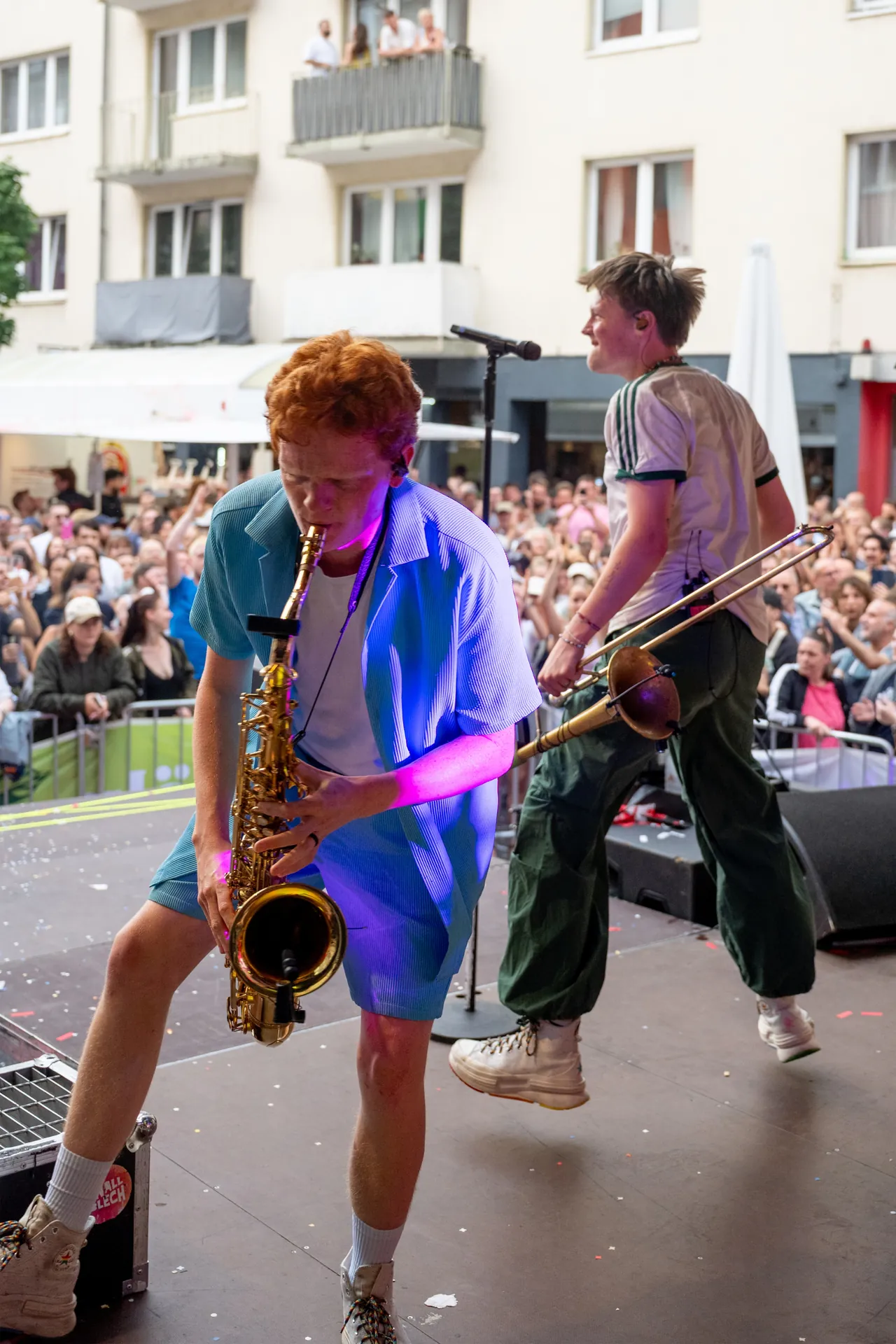 Zwei junge Musiker performen auf einer Bühne vor Publikum, einer spielt Saxofon, der andere eine Posaune.