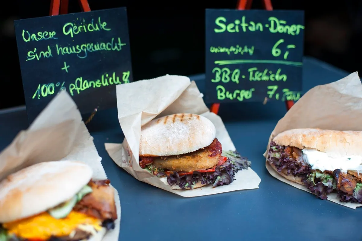 Auf einem Streetfood-Stand liegen drei vegane Gerichte mit Preisschildern zum Verkauf bereit, darunter Burger und Doner in Papier verpackt.
