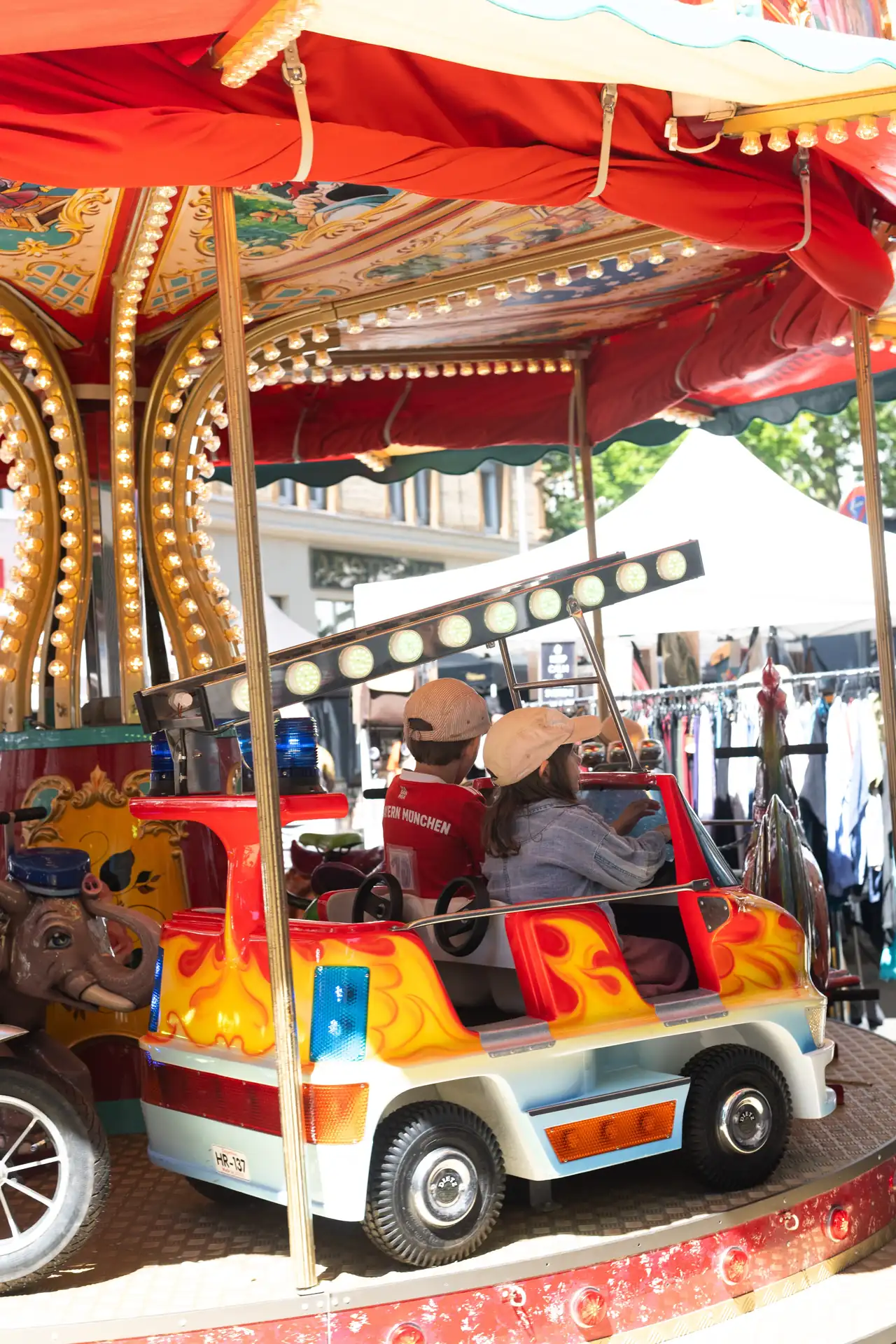 Zwei Kinder fahren in einem roten Oldtimer-ahnlichen Wagen auf einem Karussell mit roten und goldenen Verzierungen.
