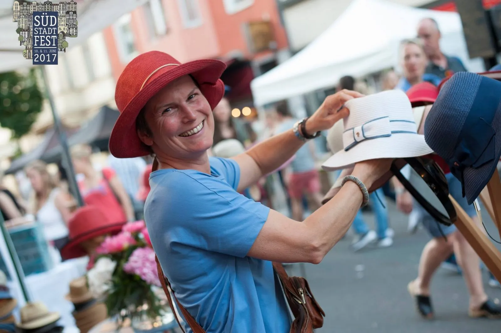 Lächelnde Frau mit rotem Hut probiert Hüte an einem Marktstand