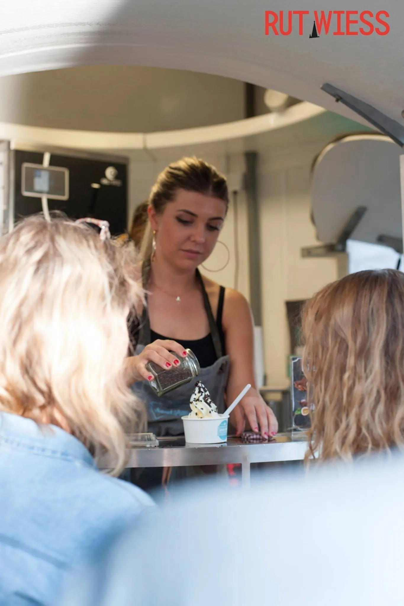 Freundliche Verkäuferin verfeinert ein Eis mit Streuseln für Kundinnen an einem sonnigen Tag