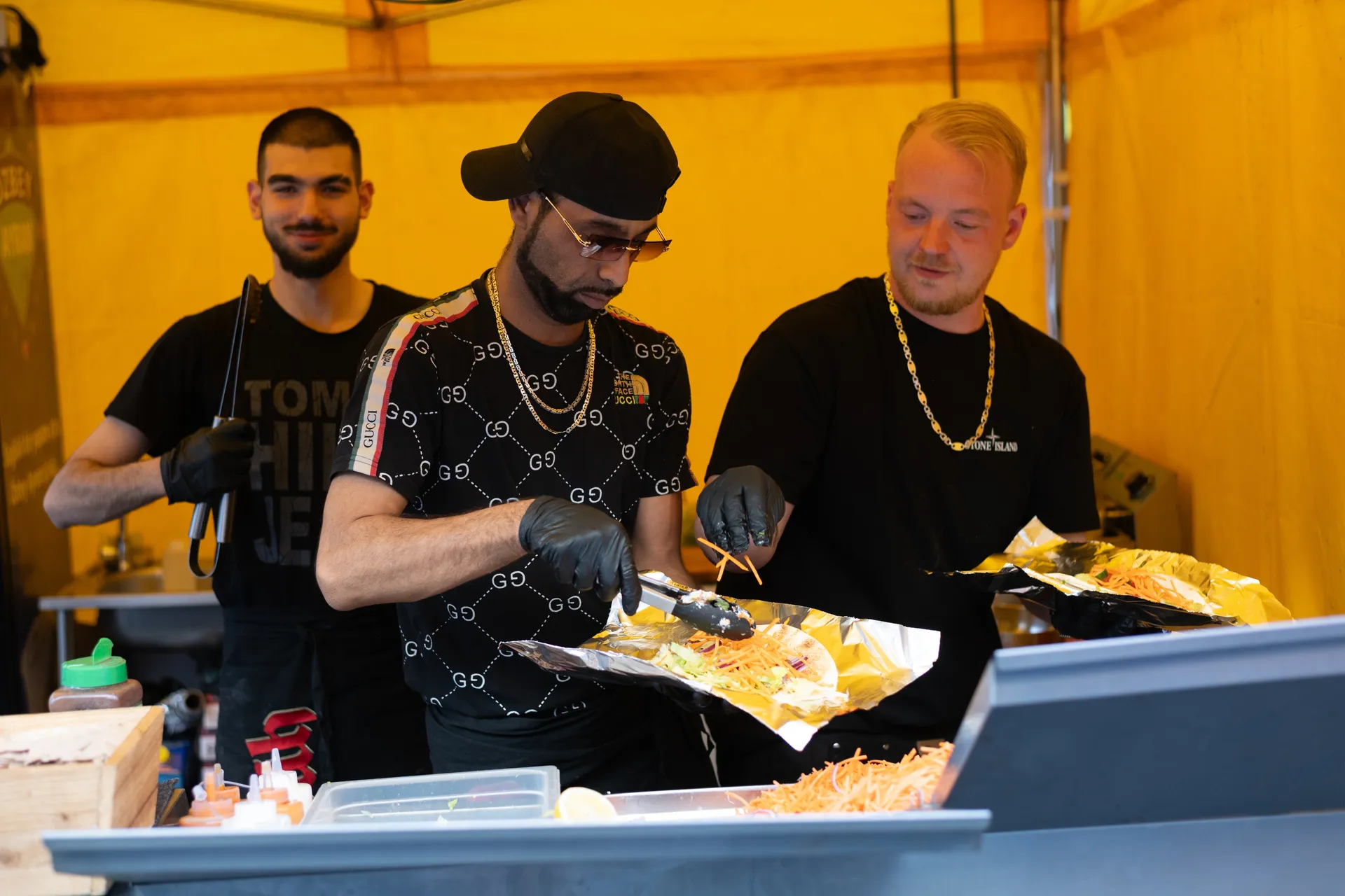 Drei Männer in schwarzer Kleidung bereiten Streetfood an einem Imbissstand unter einem gelben Zelt zu